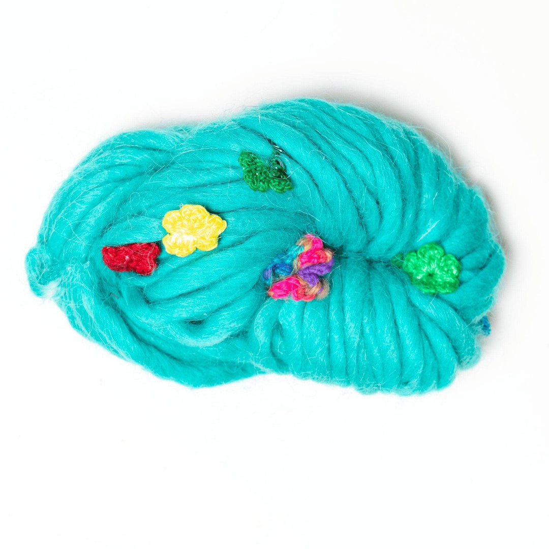 one skein of blue/green wool yarn with multicolor handmade silk flowers attached in front of a white background. 