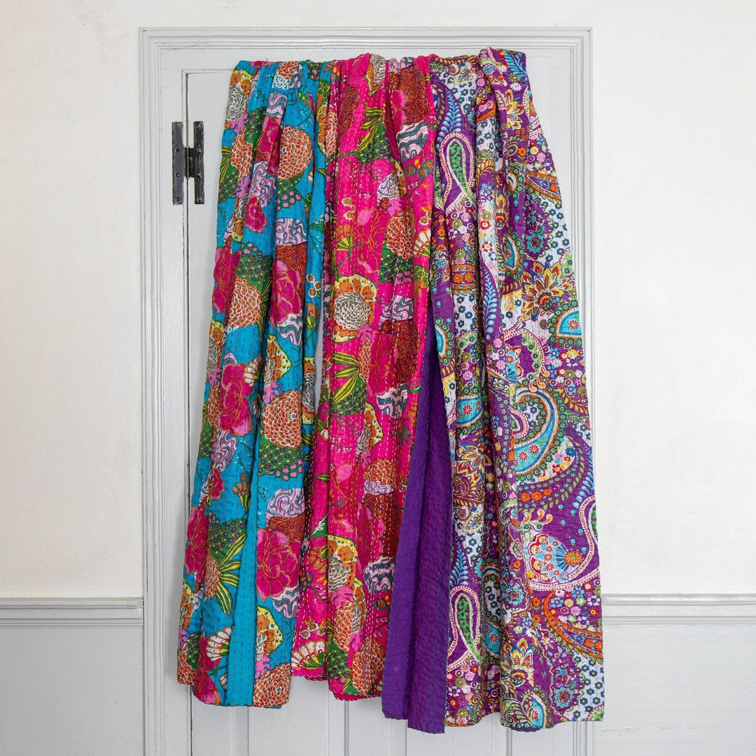 3 kantha quilts draped over the back of a white door, with a white wall surrounding. From left to right is blue, pink, and purple. 