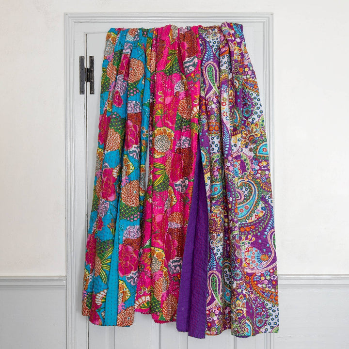 3 kantha quilts draped over the back of a white door, with a white wall surrounding. From left to right is blue, pink, and purple. 