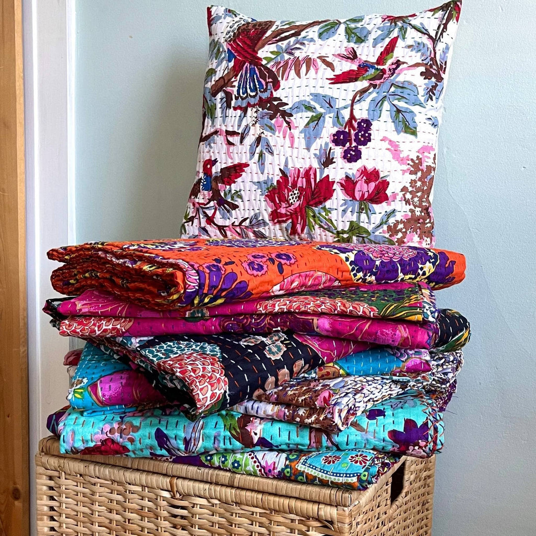 Stack of assorted  kantha quilt blankets on top of a wooden hamper with a vibrant rainforest  kantha pillow on top. 