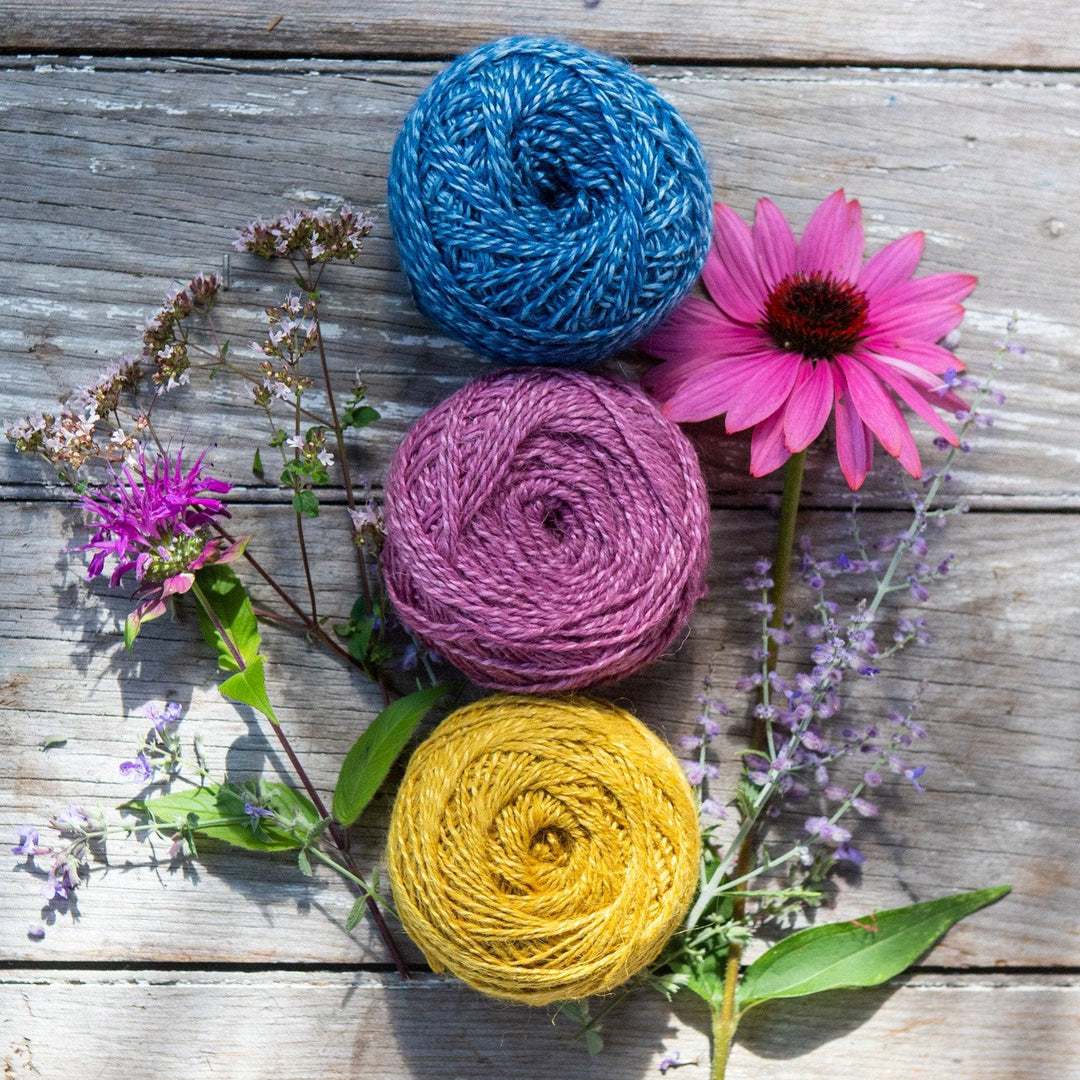 3 caked of herbal dyed silky wool plied yarn in front of a distressed wood background with several sprigs of wild flowers peeking out around the yarn. From top to bottom, blue, pink, and yellow.