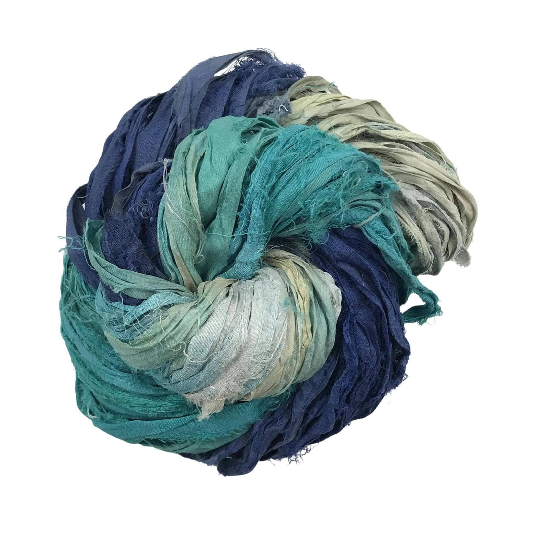 Single skein of sari silk ribbon yarn (darn blue, light blue, neutral) in front of a white background