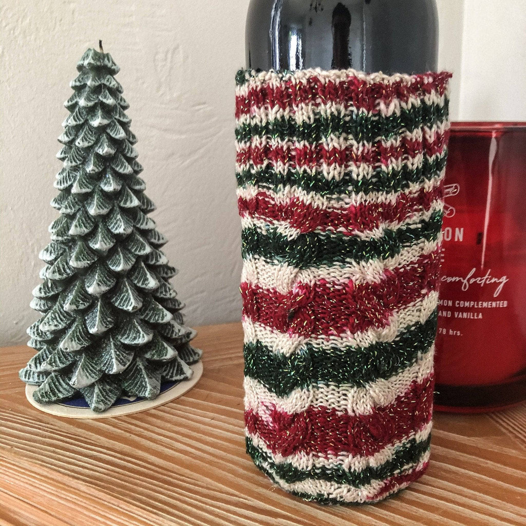 Red, green and white bottle holder with a wine bottle inside of it with a christmas tree and candle in the background