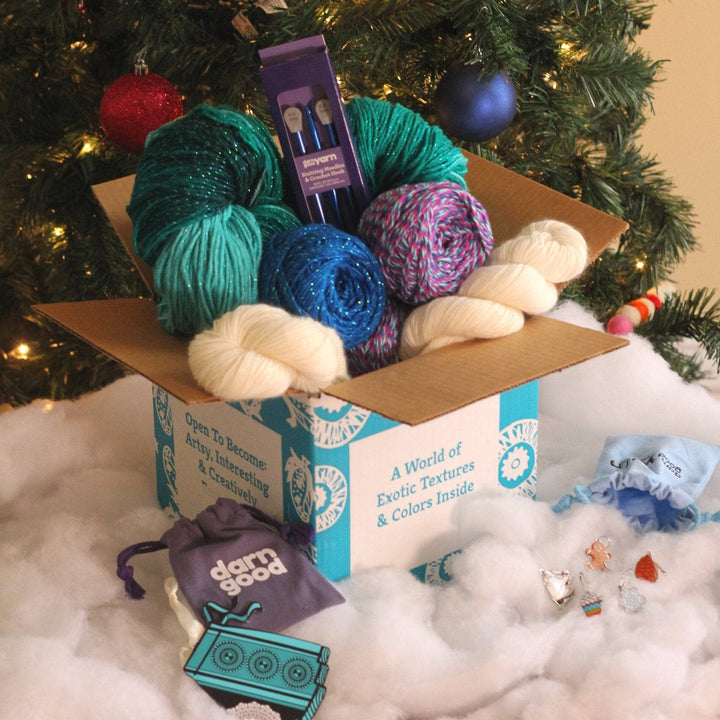 The 7th Edition Ice Queen Editors Box sitting  in front of a Christmas tree on a pile of fake snow. A skirt ornament is laying on the right side and winter themed stitch markers are on the left. Winter colored yarn is coming out of the top of the box.