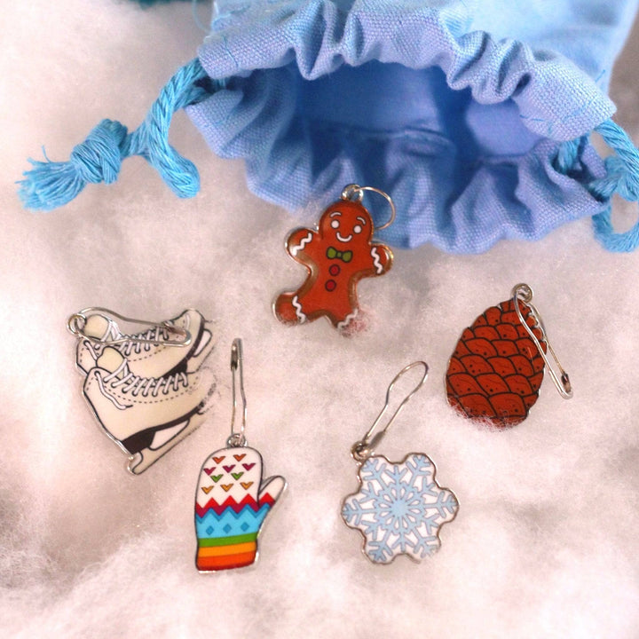 Winter themed stitch markers laying in a pile of fake snow. There's a pair of ice skates, a pine cone, a gingerbread man, a rainbow colored mitten and a snowflake.