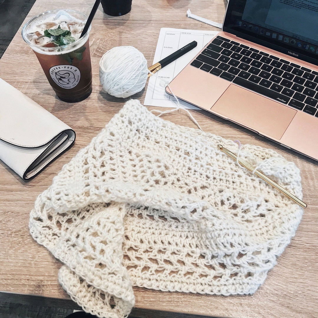 The Calla Cowl Bandana sitting on a wooden desk with iced coffee, a laptop, a wallet, and a pen and paper