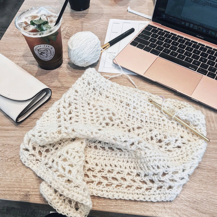 The Calla Cowl Bandana sitting on a wooden desk with iced coffee, a laptop, a wallet, and a pen and paper
