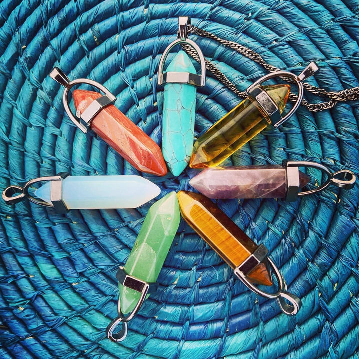 Interchangeable Chakra necklace charms arranged in a star formation upon a blue basket background. The crystals in clockwise order from the top are blue, transparent yellow, purple, solid yellow, green, light blue, and red.