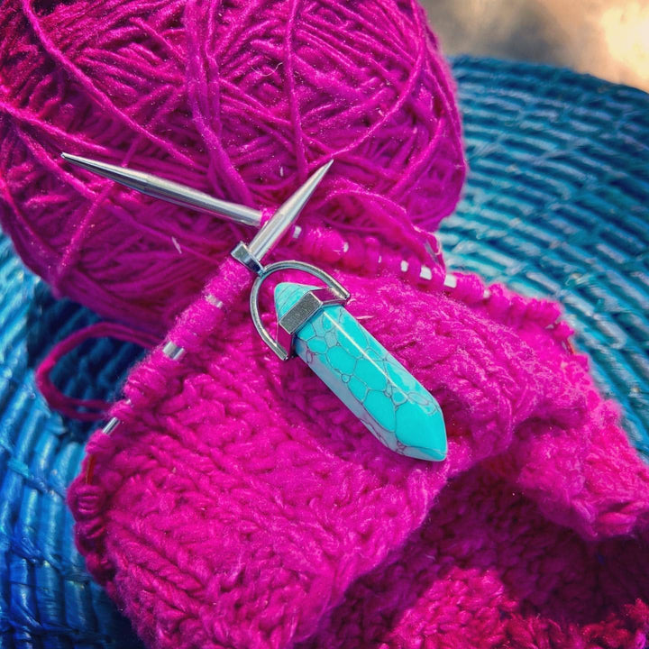 Blue chakra pendant hanging from a knitting needle with bright pink yarn on a blue basket.