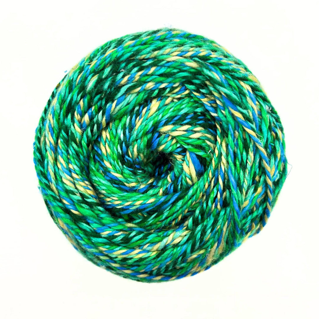 one skein of journey recycled silk yarn in the Indian Peacock colorway (variegated green, yellow, blue) in front of a white background.