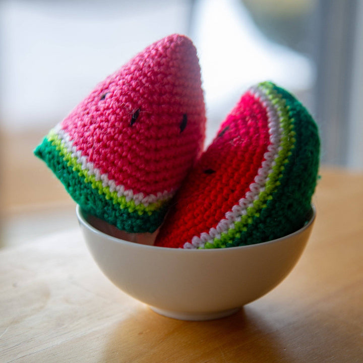 2 Crocheted Watermelon amigurumi slices sitting in a white bowl on a wooden surface