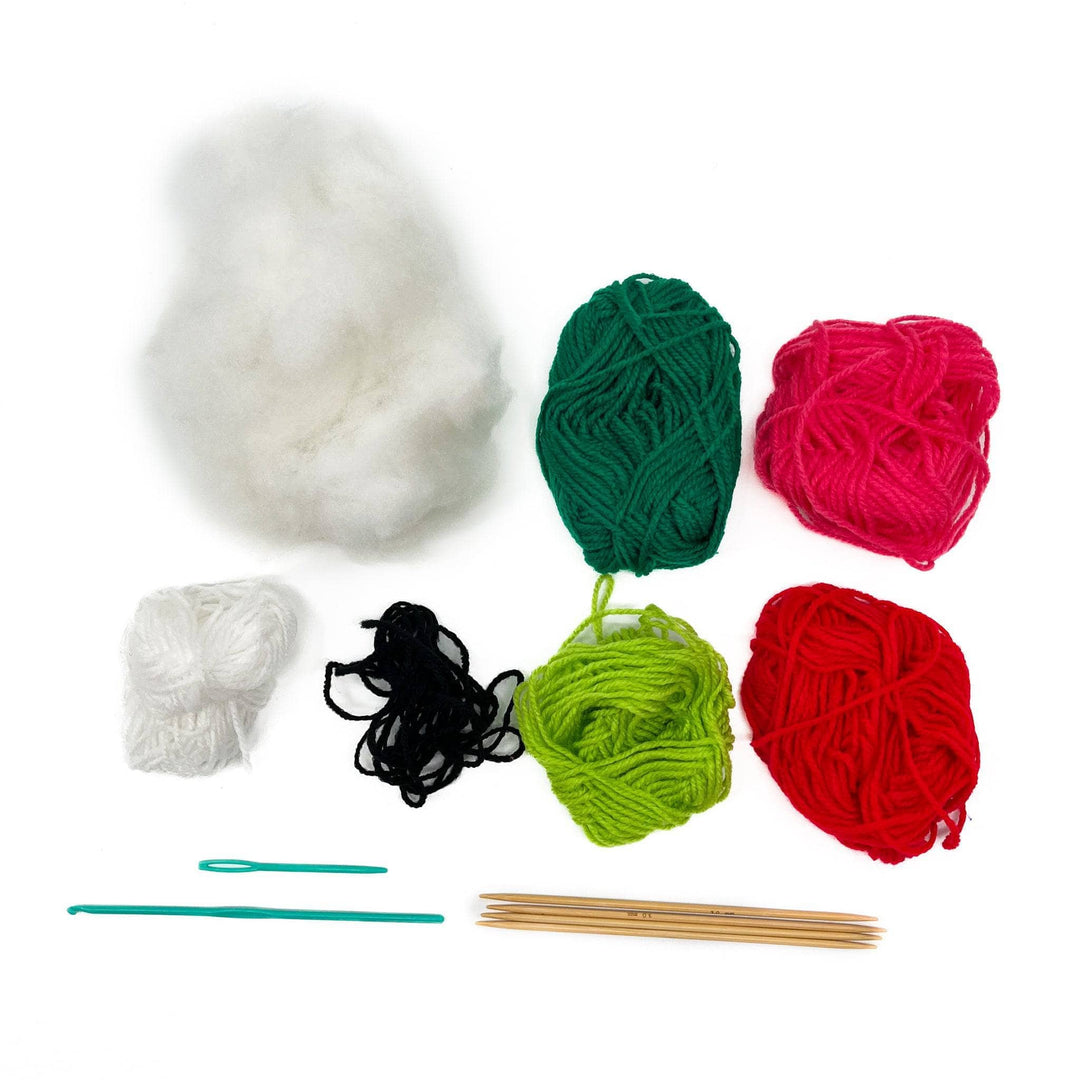 Image of all the supplies that comes in the kit, fluffy white stuffing, different colored yarns dark green, light green, pink, red, white and black. A crochet hook, four wooden knitting needles and a plastic sewing needle 