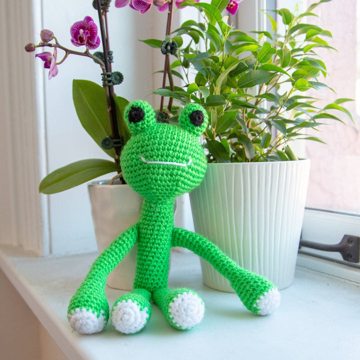 lifestyle shot of completed amigurumi frog sitting on a windowsill with potted plants in the background.