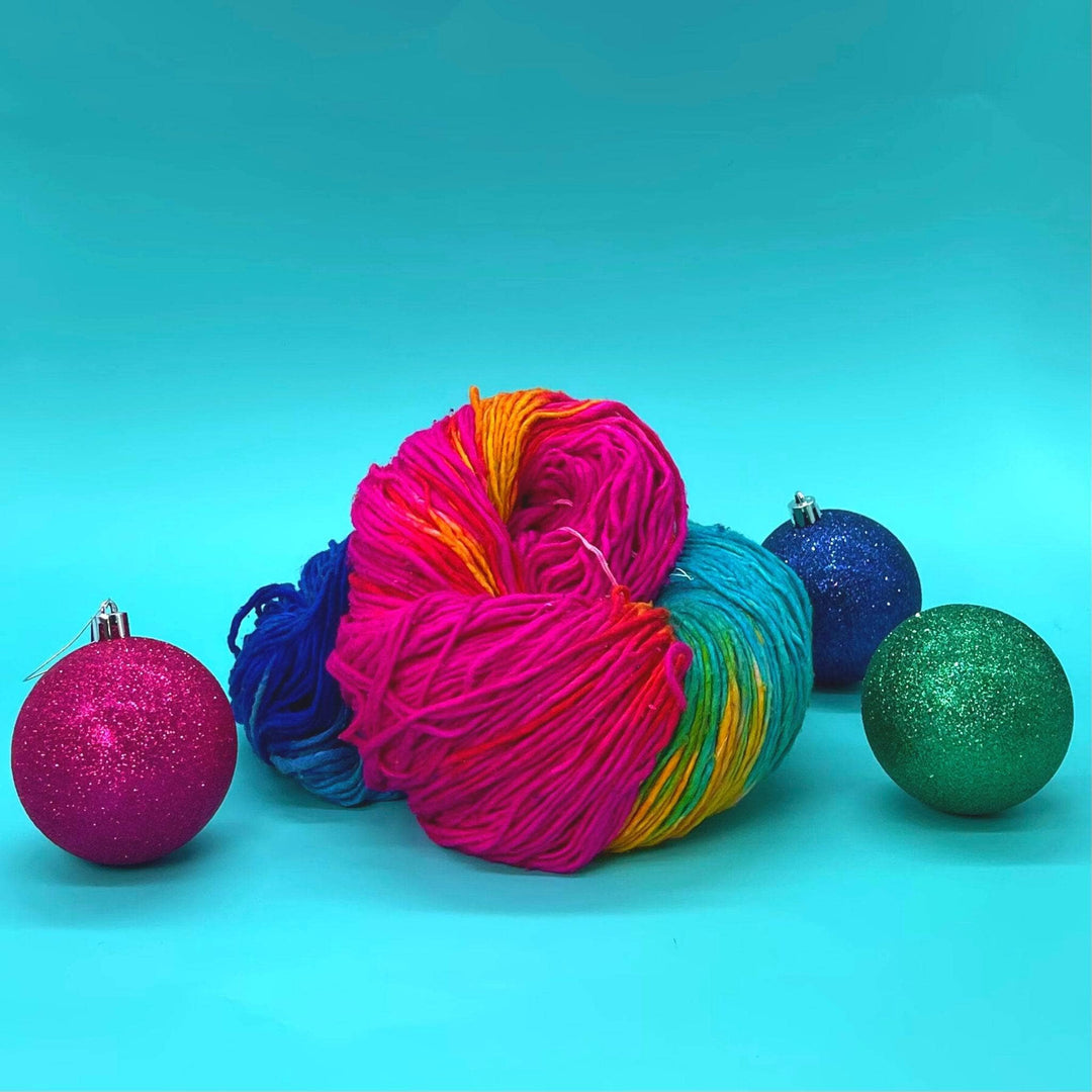 A mega skein of silk roving worsted weigh silk yarn in the colorway Vibrant Macaron. This yarn is a rainbow ombre yarn with dark to light blues, greens, pinks and oranges. The yarn is in a birds nest sitting on a blue background with the rainbow ornaments around it to show how big it its.