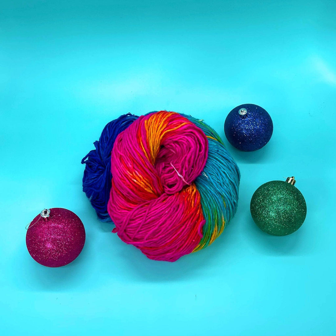 An overhead view of a mega skein of silk roving worsted weigh silk yarn in the colorway Vibrant Macaron. This yarn is a rainbow ombre yarn with dark to light blues, greens, pinks and oranges. The yarn is in a birds nest sitting on a blue background with the rainbow ornaments around it to show how big it its.