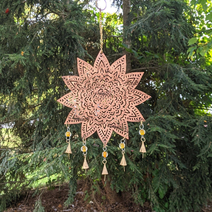 Copper plated metal mandala chime hanging with greenery in the background.