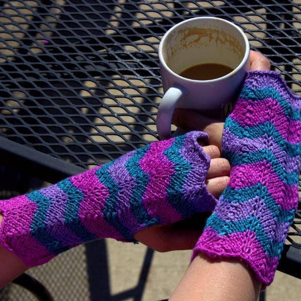 close up of hands wearing  purple green and pink mittens and holding a coffee mug