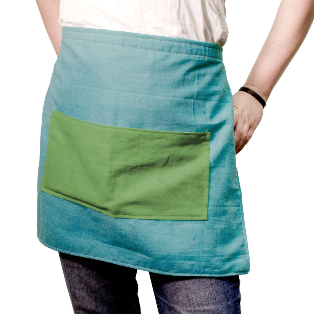 model wearing a blue and green apron with front pocket in front of a white background.
