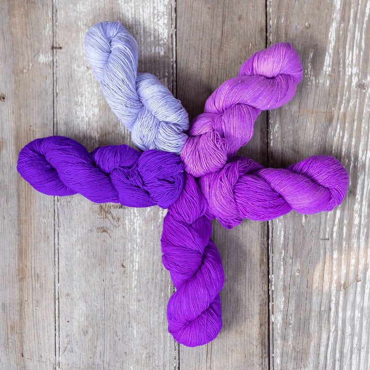 5 skeins of sport weight silk in the colorway purples arranged in a star formation in front of a wood background.