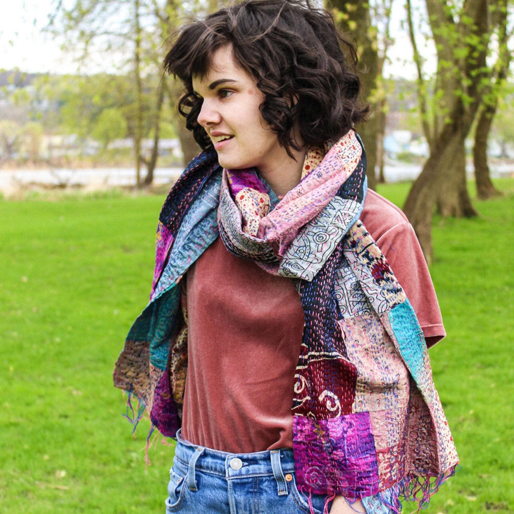 Model wearing one of a kind kantha scarf, patchwork with many colors, worn with blue jeans and a pink tee shirt. Model is outside with green grass and trees in the background.