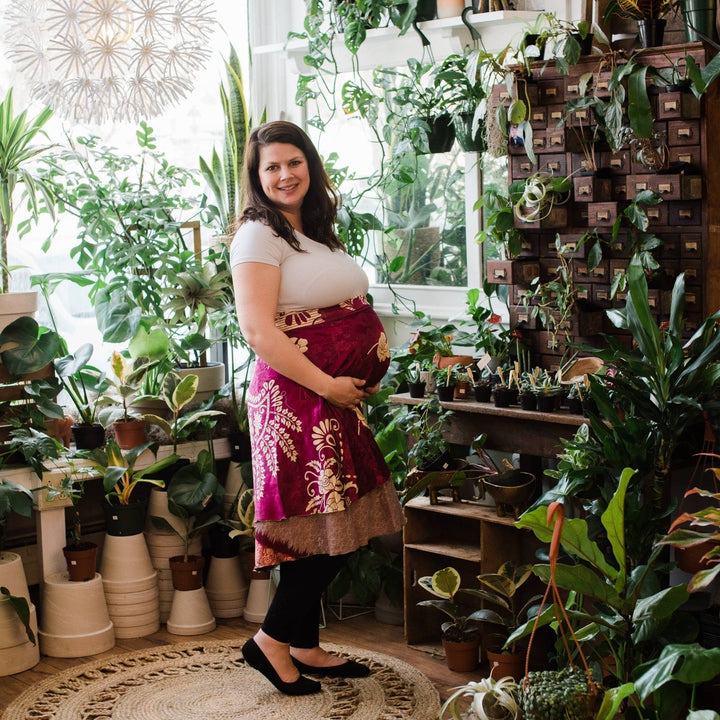 Pregnant model is wearing dark pink tea length sari wrap skirt with potted plant background. 