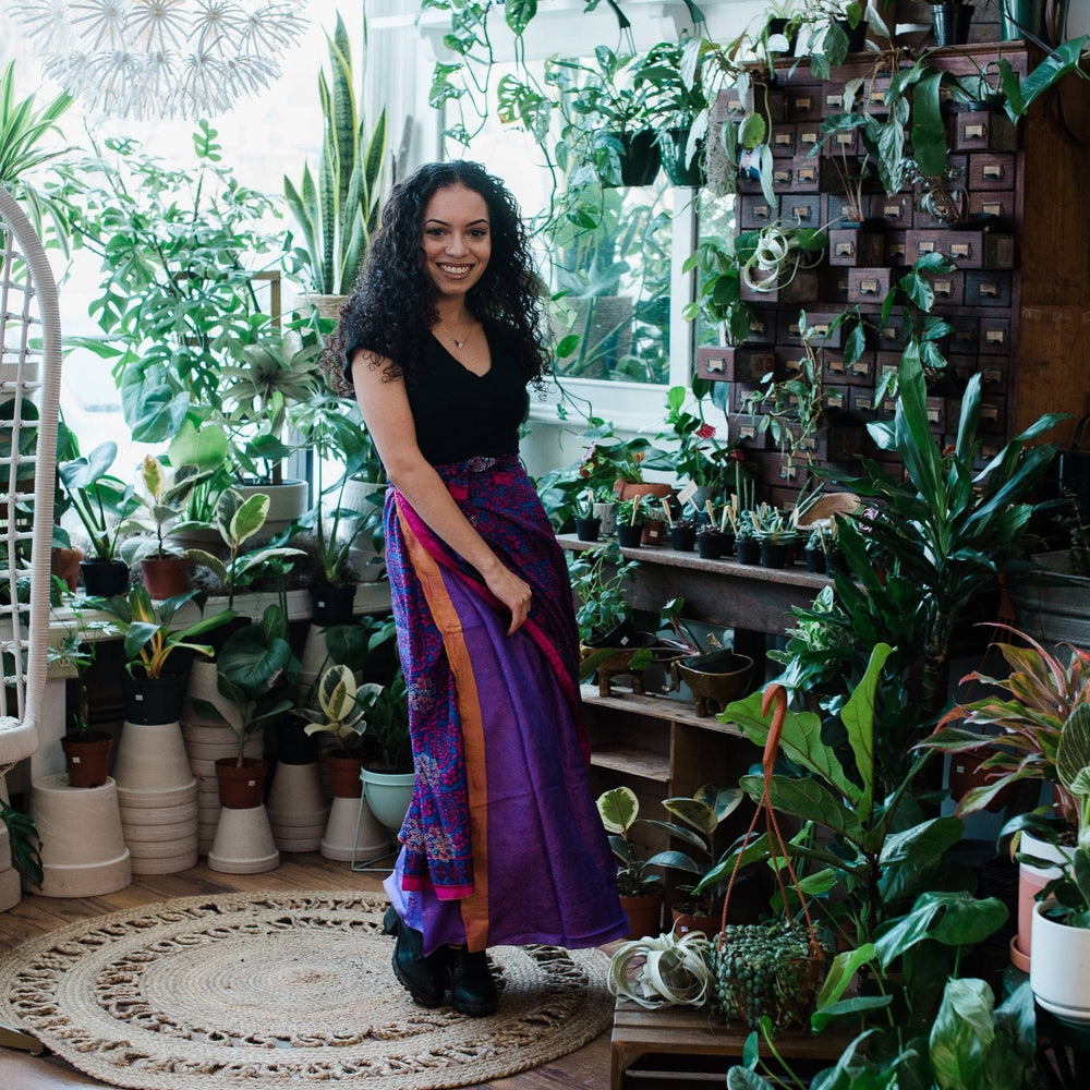 Model is wearing one of a kind purple, orange and pink long wrap sari skirt with potted plants background. 