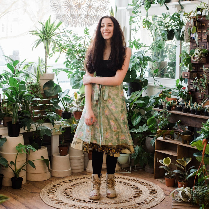 Model wearing green and gold tea length sari wrap skirt with potted plants in the background.  
