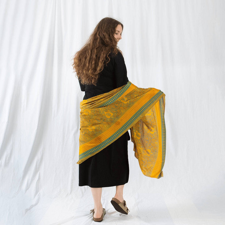 Model is standing in front of a white backdrop with a yellow one of a kind sari silk scarf wrapped around their waist. 