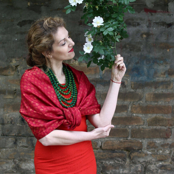 model wearing one of a kind sari short scarf in red while smelling a dangling vine of flowers with brick wall in the background.