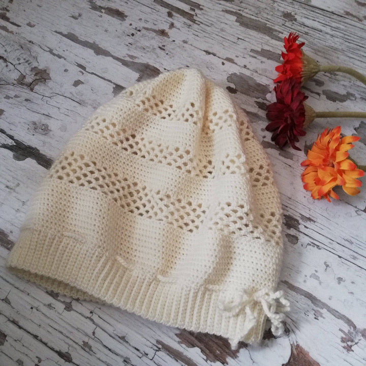 white crochet beanie with lace stitching laying on a wood surface with distressed white paint and 3 flowers beside.