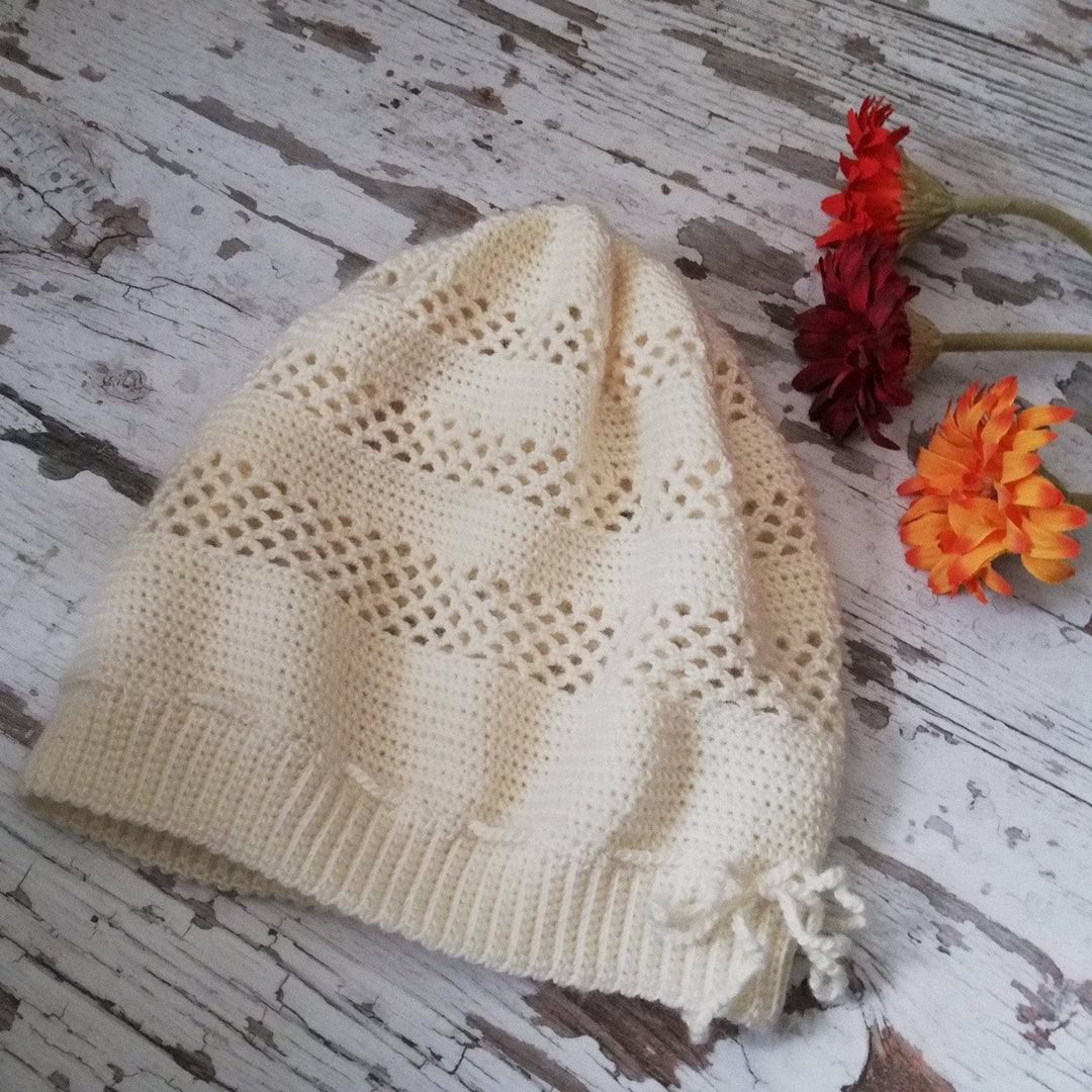 white crochet beanie with lace stitching laying on a wood surface with distressed white paint and 3 flowers beside.