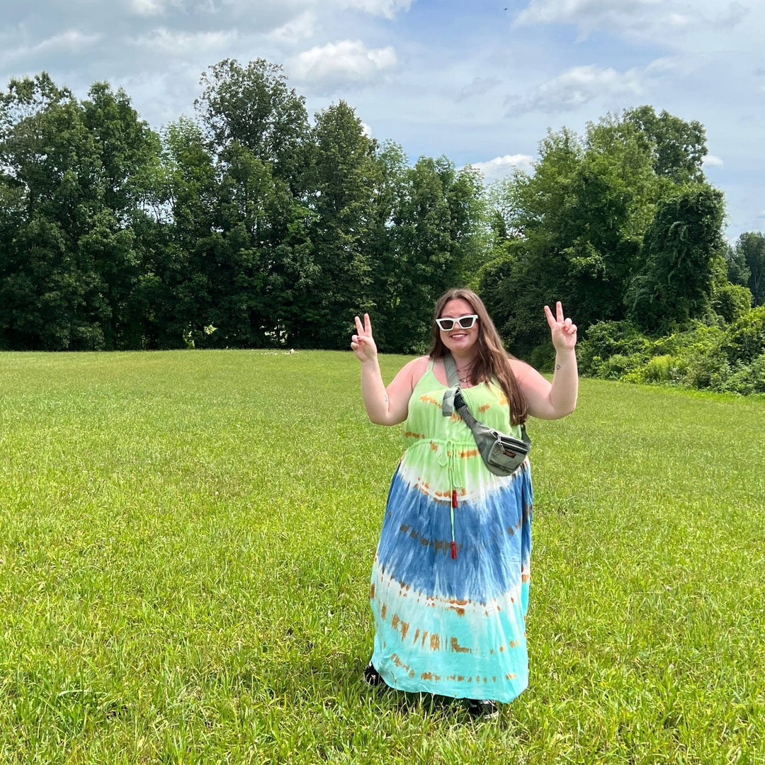Model is standing in a field holding up a peace signs with both hands wearing the blue breeze tie dye maxi dress. 