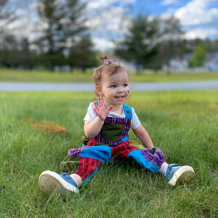Toddler wearing purple, blue and green patchwork play pants while sitting in the grass. 