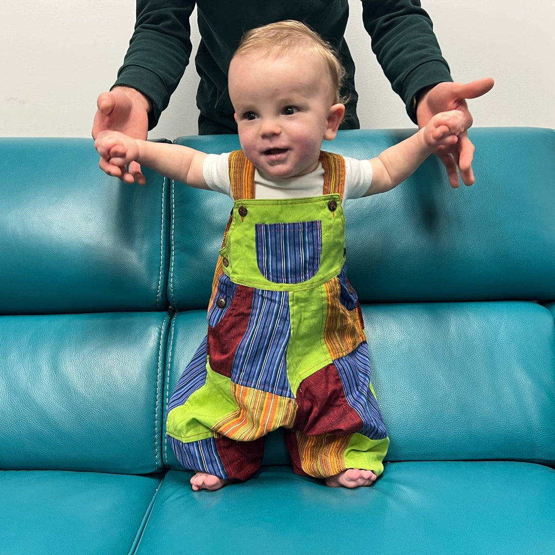 Baby wearing green, blue, orange and red patchwork overalls while standing on a turquoise couch holding an adult's hands for balance.  