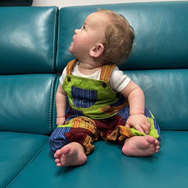 Baby wearing patchwork overalls in green, blue, orange and red while sitting on a turquoise couch.