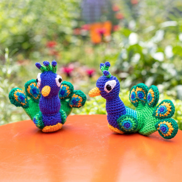 Knit (left) and crochet (right) peacocks sitting next to each other on a n orange table with greenery in the background.