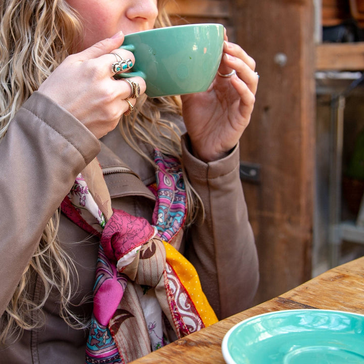 Model is out to eat sipping a coffee wearing a pink and yellow medley scarf. 