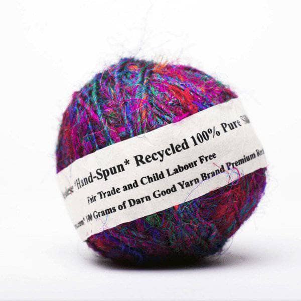 close up of a multicolored cake of yarn with a white tag which reads "Hand-spun 100% Recycled. Fair trade, and child labor free. 100 grams of Darn Good Yarn brand premium recycled sari silk yarn."