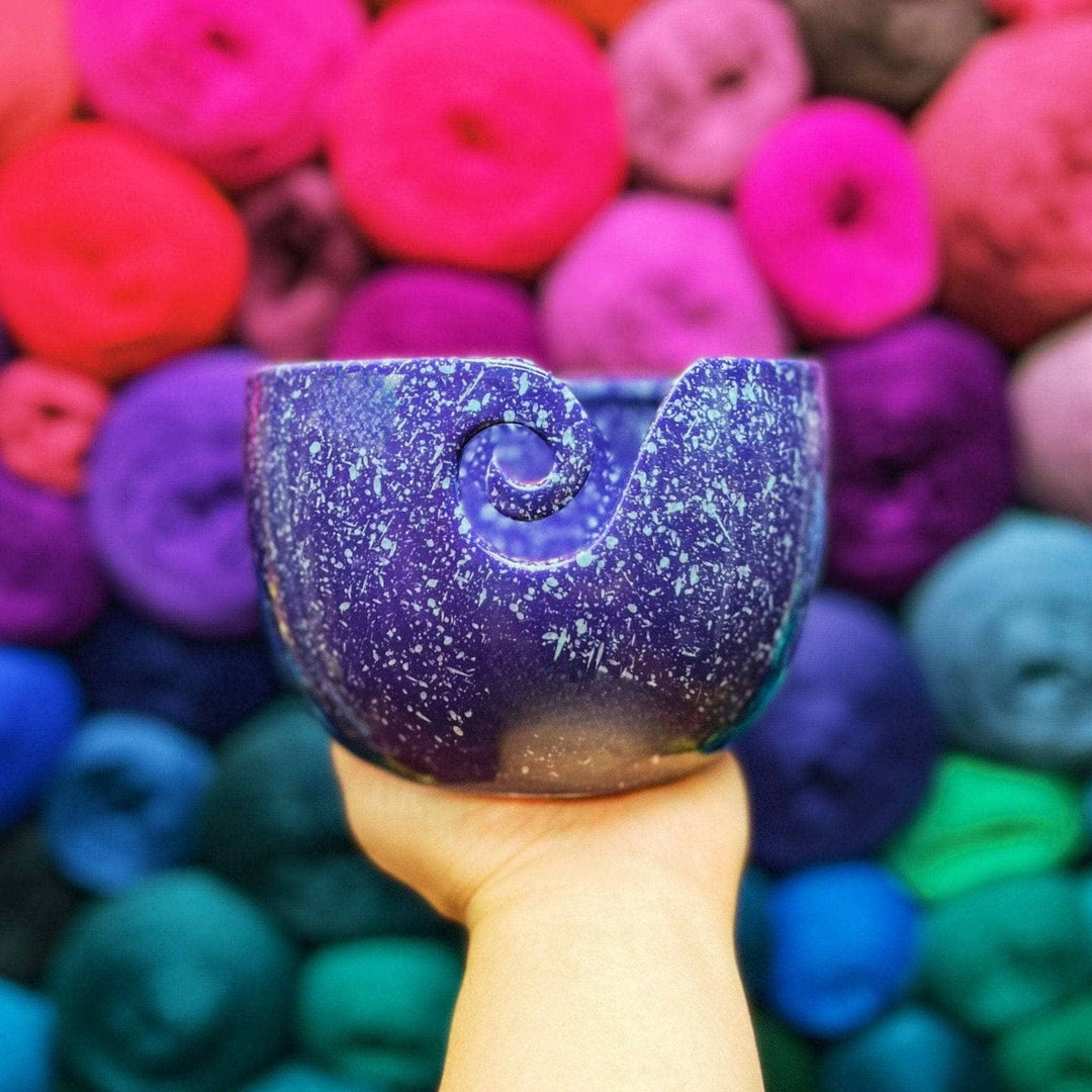 Hand holding up a purple ceramic yarn bowl with white speckles with multicolored yarn in the background.