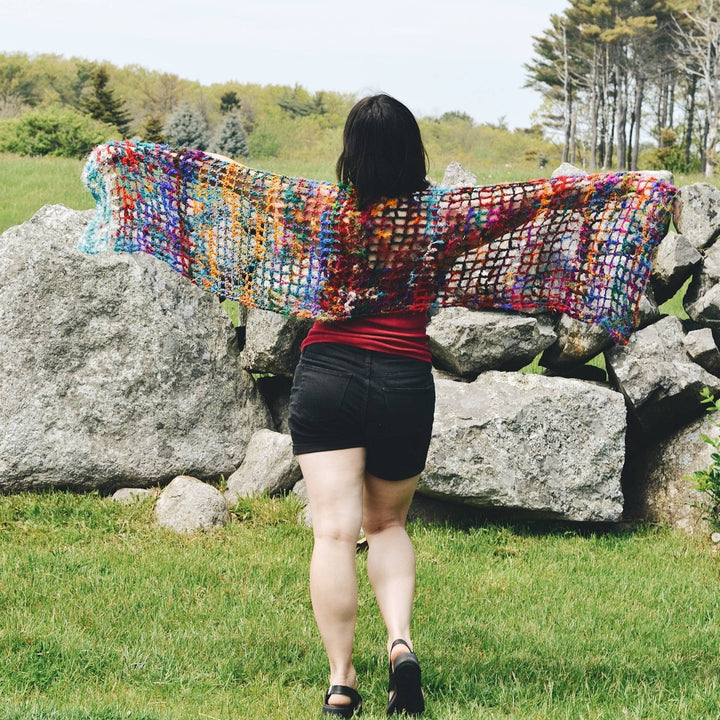 Back view of model wearing quadramesh shawl stretched out with rocks and greenery in the background.