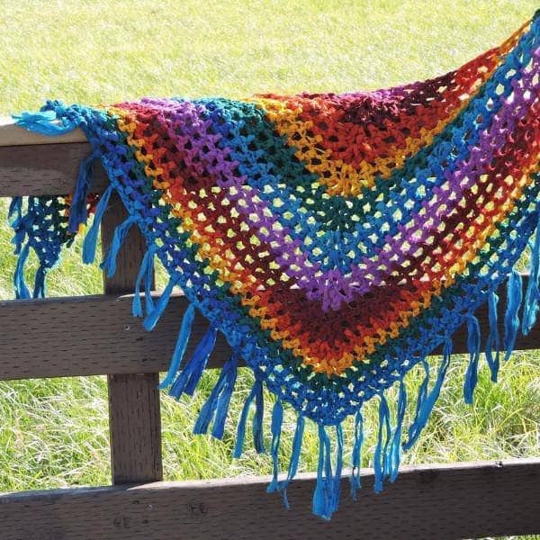 Rainbow Shawl laying over a brown fence with a field in the background