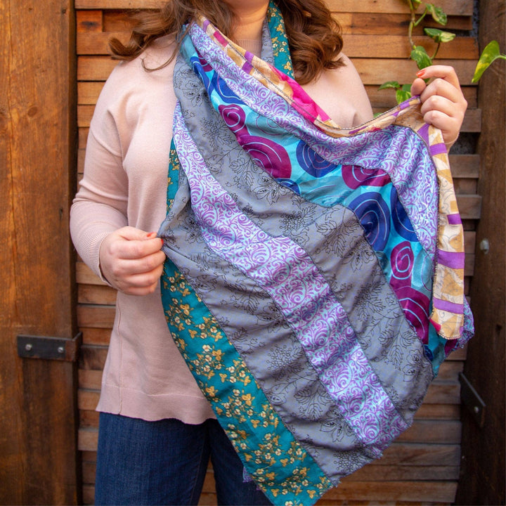 Model wearind sari silk medley scarf (blue multicolor) and holding it outstretched with a brown wood wall in the background.