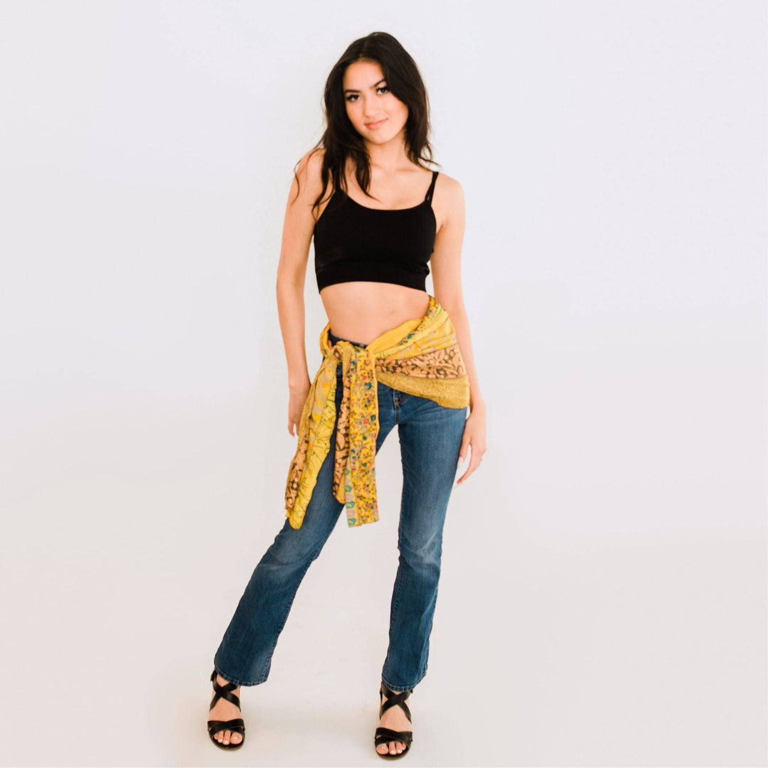 A woman standing in front of a white back drop, wearing a cropped black tank top and blue jeans. She's added a mustard yellow sari medley around her waist like a sarong, adding a pop of color to her look.