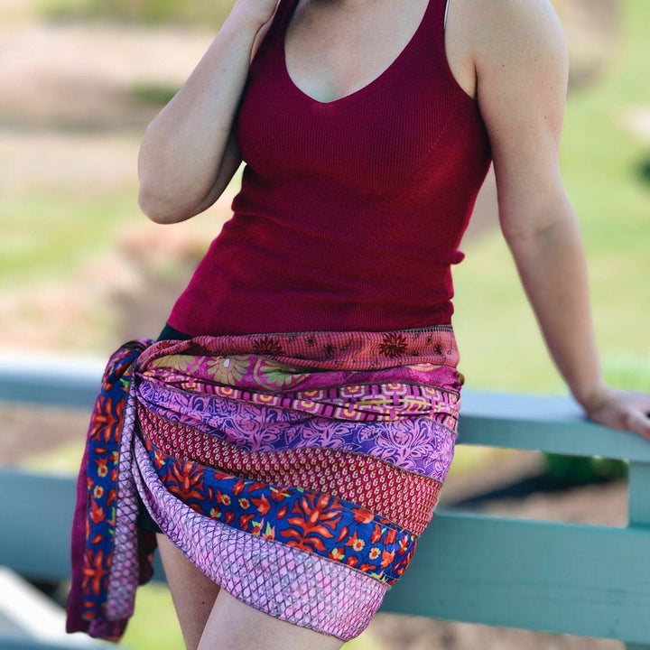 A woman standing on a porch outside, she's wearing jean shorts and a maroon tank top. She's wearing a medley around her waist like a sarong. The medley is made from 7 strips of reclaimed sari material in pinks, reds and maroons.