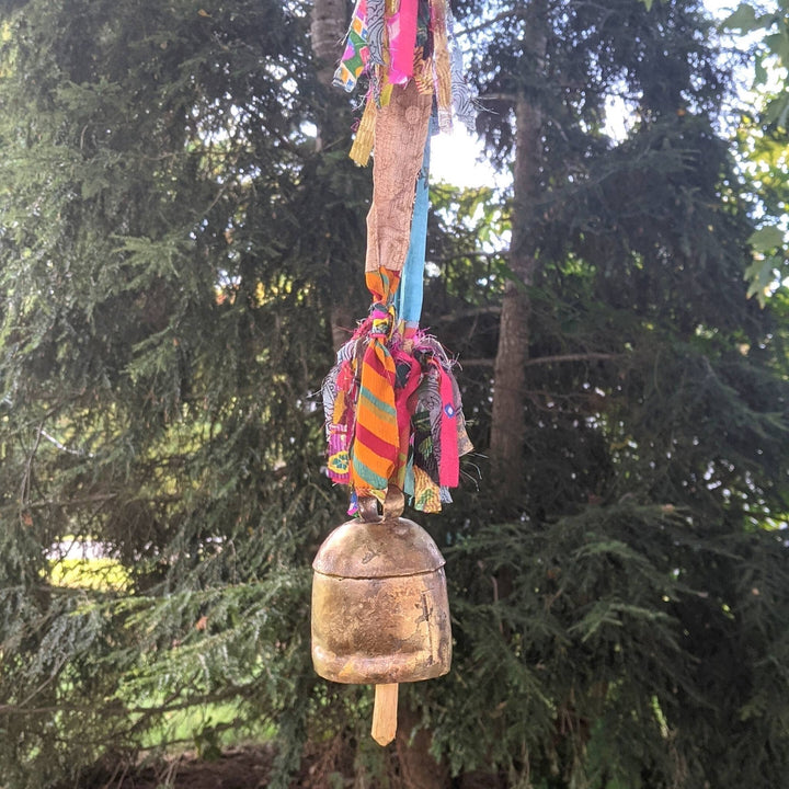 Brassy colored bell with wooden clapper, hanging from a colorful sari ribbon with 4 fringe tassels in with trees in the background.
