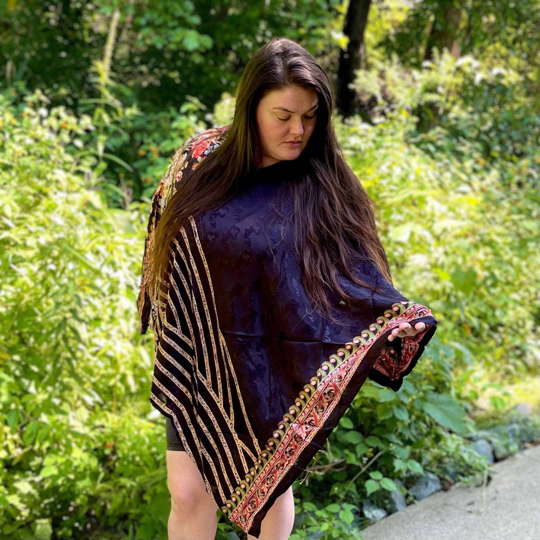 A woman standing in a forest wearing a black poncho made from reclaimed sari material. This sari poncho has gold stripes along the edge and delicate floral patterns on the shoulder and hood.