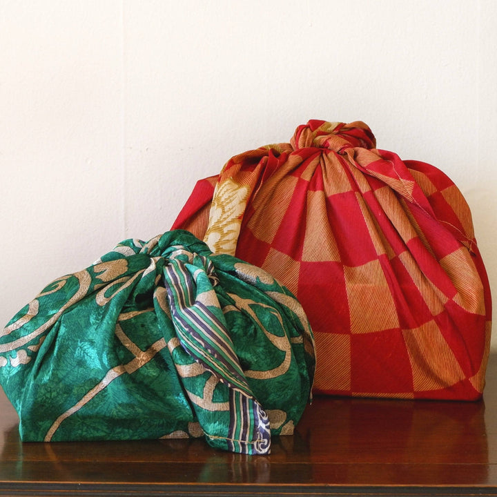 Two square presents wrapped in different sized furoshiki gift wrapping. The larger is a maroon and gold checkered sari and the smaller is an emerald with gold details. The furoshiki gift wrap is made from reclaimed sari.