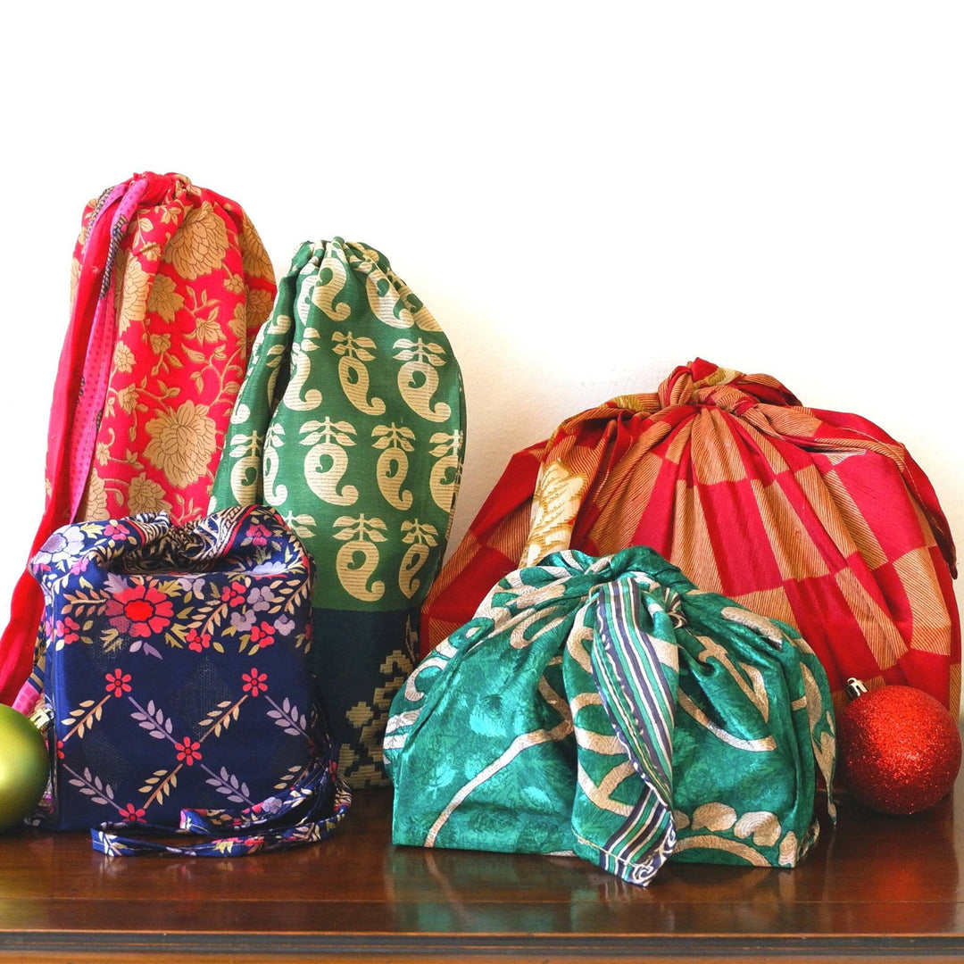5 presents sitting on a dark wood floor on a white background. There are 3 presents wrapped in 3 various sized draw string bags, 2 wrapped in various sized furoshiki wrapping. Each present features a different pattern and color as they're all made from reclaimed sari.