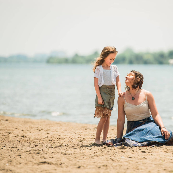 Two models sitting on the beach. The adult model sits in the sand wearing a blue sari wrap skirt looking up at the child model standing next to them wearing a green mini sari wrap skirt. 