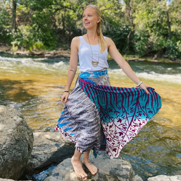 Model is standing on a rock by a stream wearing a tea length sari wrap skirt in blue, purple and sliver. 